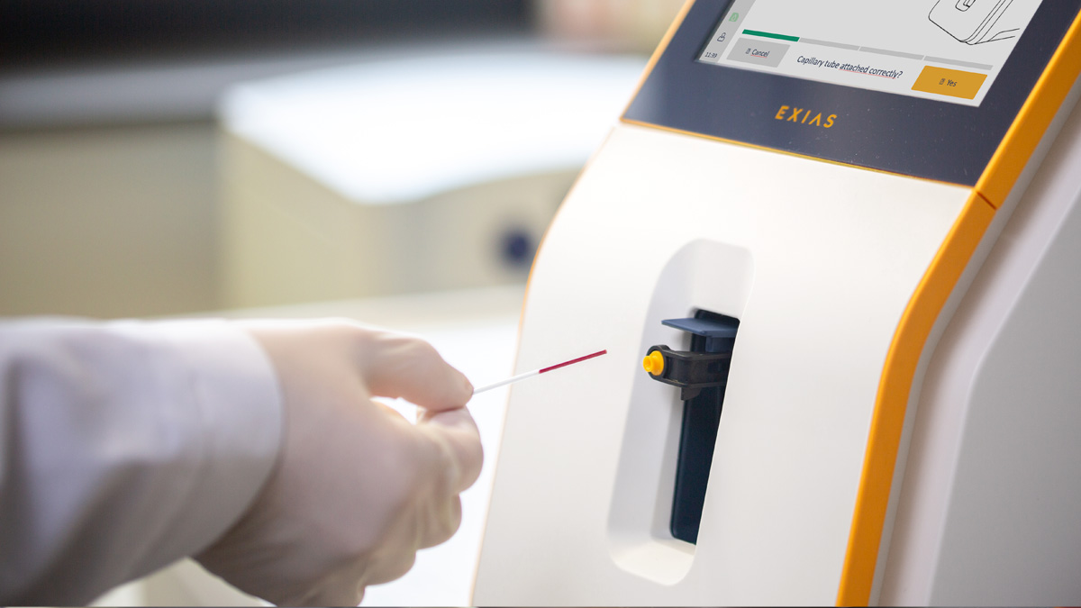Hansket hånd som setter et blodprøverør inn i en medisinsk analyseapparat med en skjerm.