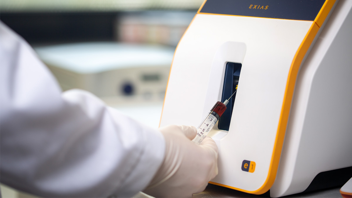 Hansket hånd setter en blodprøvesprøyte inn i en medisinsk analysemaskin merket "EXIAS" for testing.