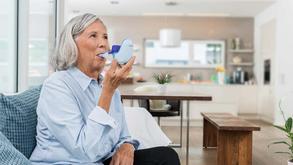 Eldre kvinne som sitter i en sofa og bruker en blå åndedrettsinhalator i et lyst, moderne kjøkken.
