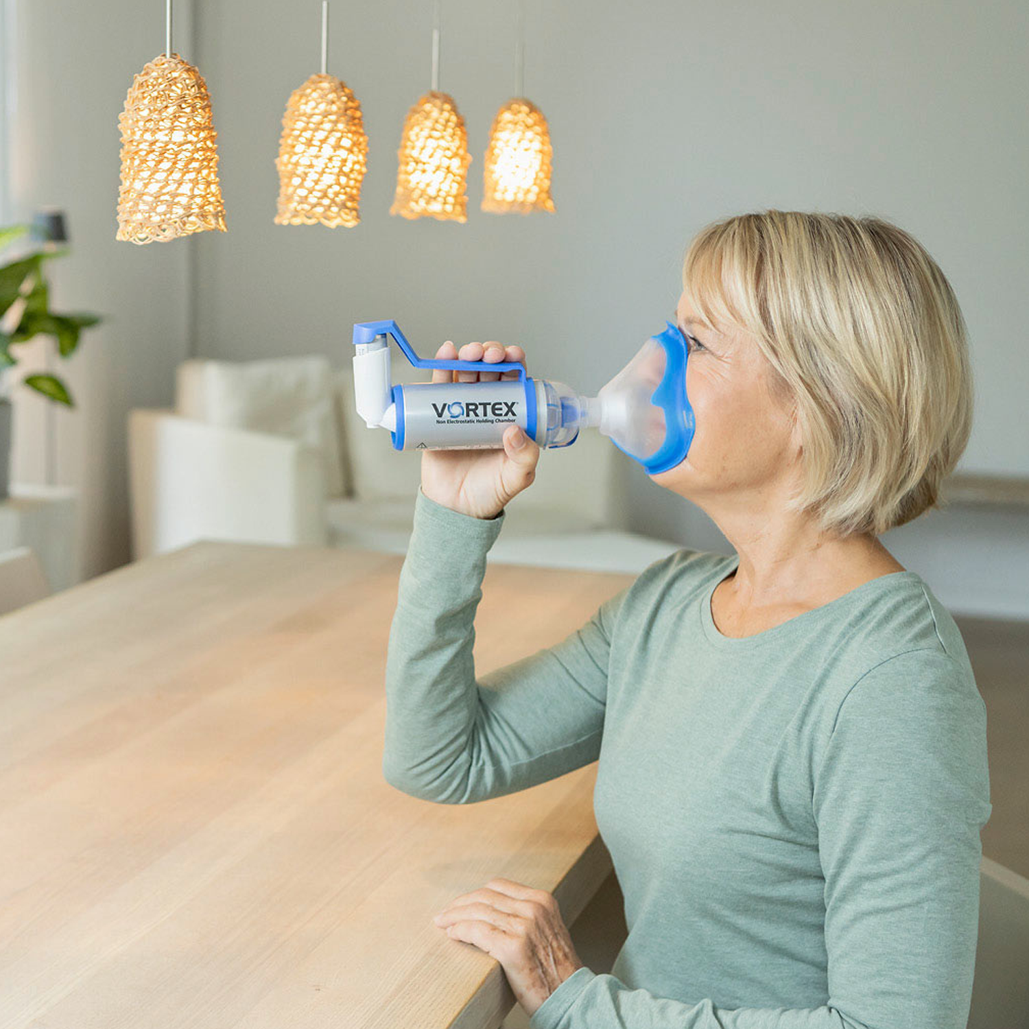 Kvinne som bruker en inhalator med forstøver på et bord i et godt opplyst, moderne rom med hengende lys.