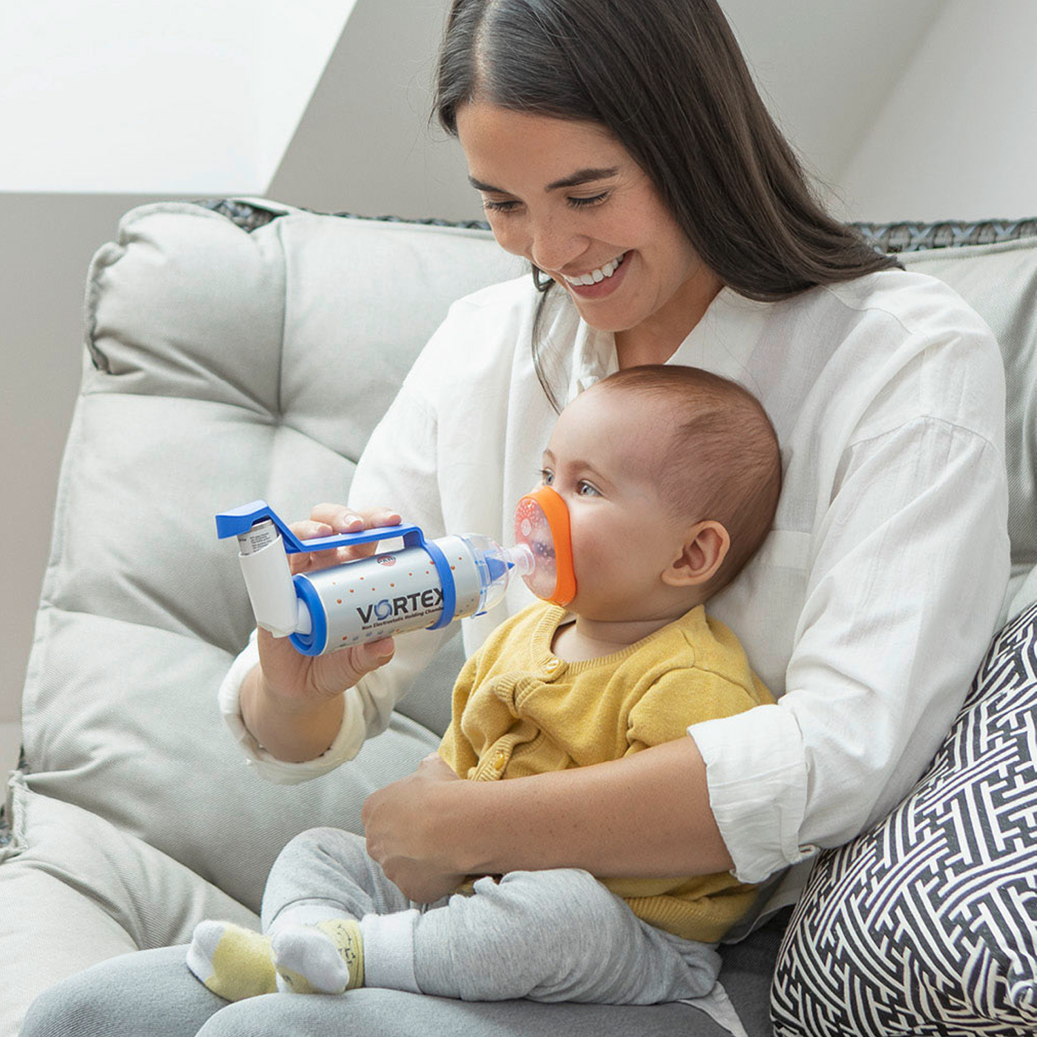 Kvinne som smiler mens hun gir en baby en inhalator med maske, sittende på en sofa.