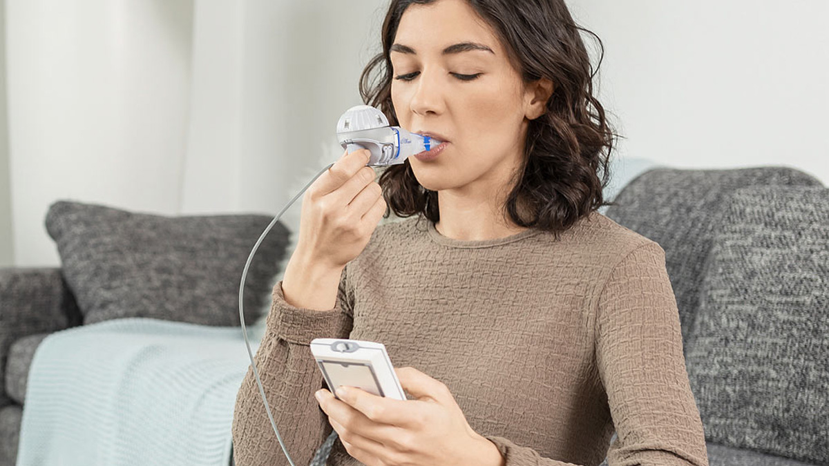 Kvinne som bruker en håndholdt respiratorisk enhet mens hun sitter på en sofa, og holder en tilkoblet mobil enhet.