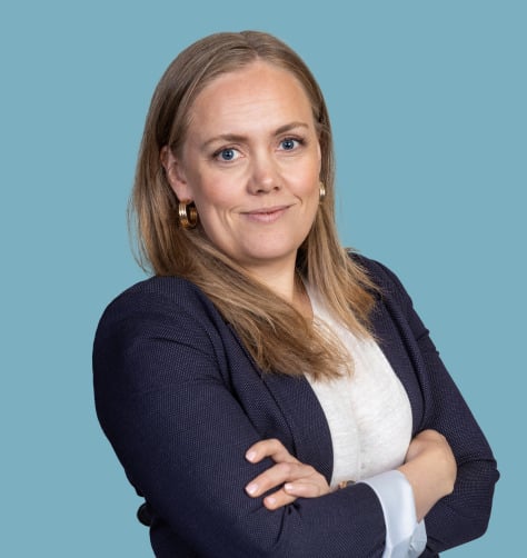 Kvinne med langt blondt hår i en marineblå blazer og hvit skjorte, med armene krysset, mot en blå bakgrunn.